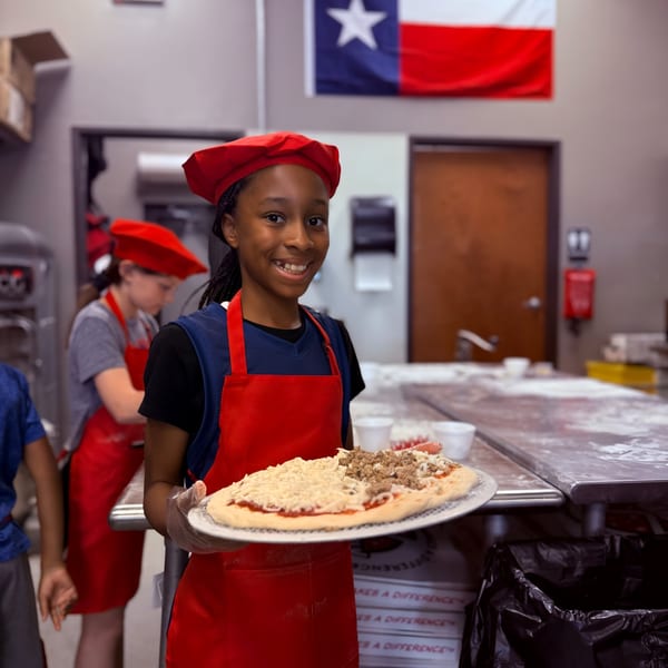A smiling girl in a red chef's hat and apron holds an uncooked pizza with cheese and meat.