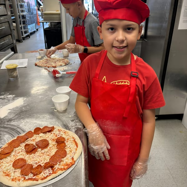 Two young boys in red chef hats and aprons making pizzas in a commercial kitchen.