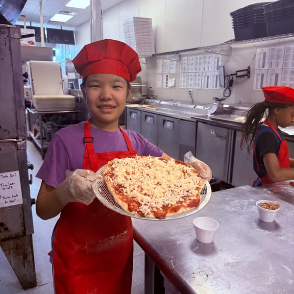 A smiling child in a red chef's hat and apron holds a pizza with sauce and cheese.