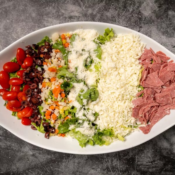 A white oval platter filled with a vibrant salad, featuring tomatoes, olives, cheese, and deli meat.