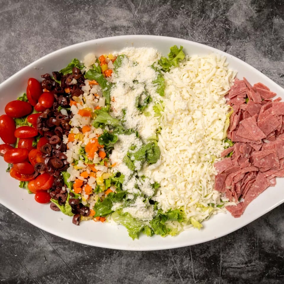 A white oval platter filled with a vibrant salad, featuring tomatoes, olives, cheese, and deli meat.
