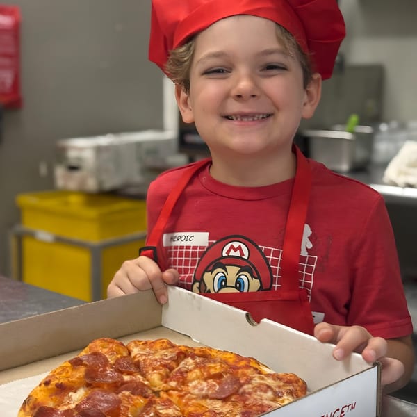 A smiling boy in a red chef's hat and Mario shirt holds an open pizza box with a pepperoni pizza.