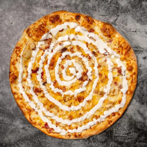 Round pizza with melted cheese, bacon, chicken, and a spiral of white sauce on a dark surface.