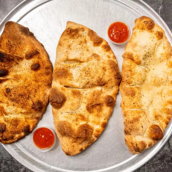 Three golden-brown calzones with two small cups of marinara sauce on a metal pizza pan.