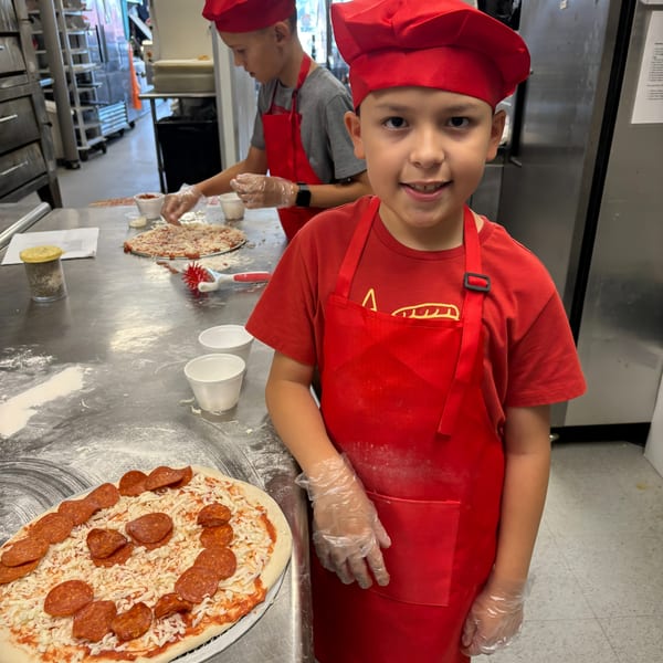 Two young boys in red chef hats and aprons making pizzas in a commercial kitchen.