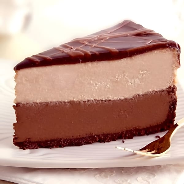 Slice of chocolate cheesecake with a light brown layer, topped with dark chocolate glaze and drizzle.