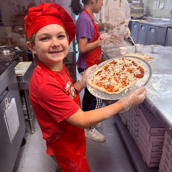 A smiling child in a red chef's hat and apron holds a pizza with sauce and cheese.