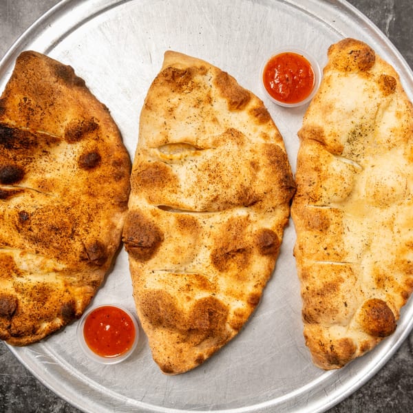 Three golden-brown calzones with two small containers of marinara sauce on a metal pizza pan.